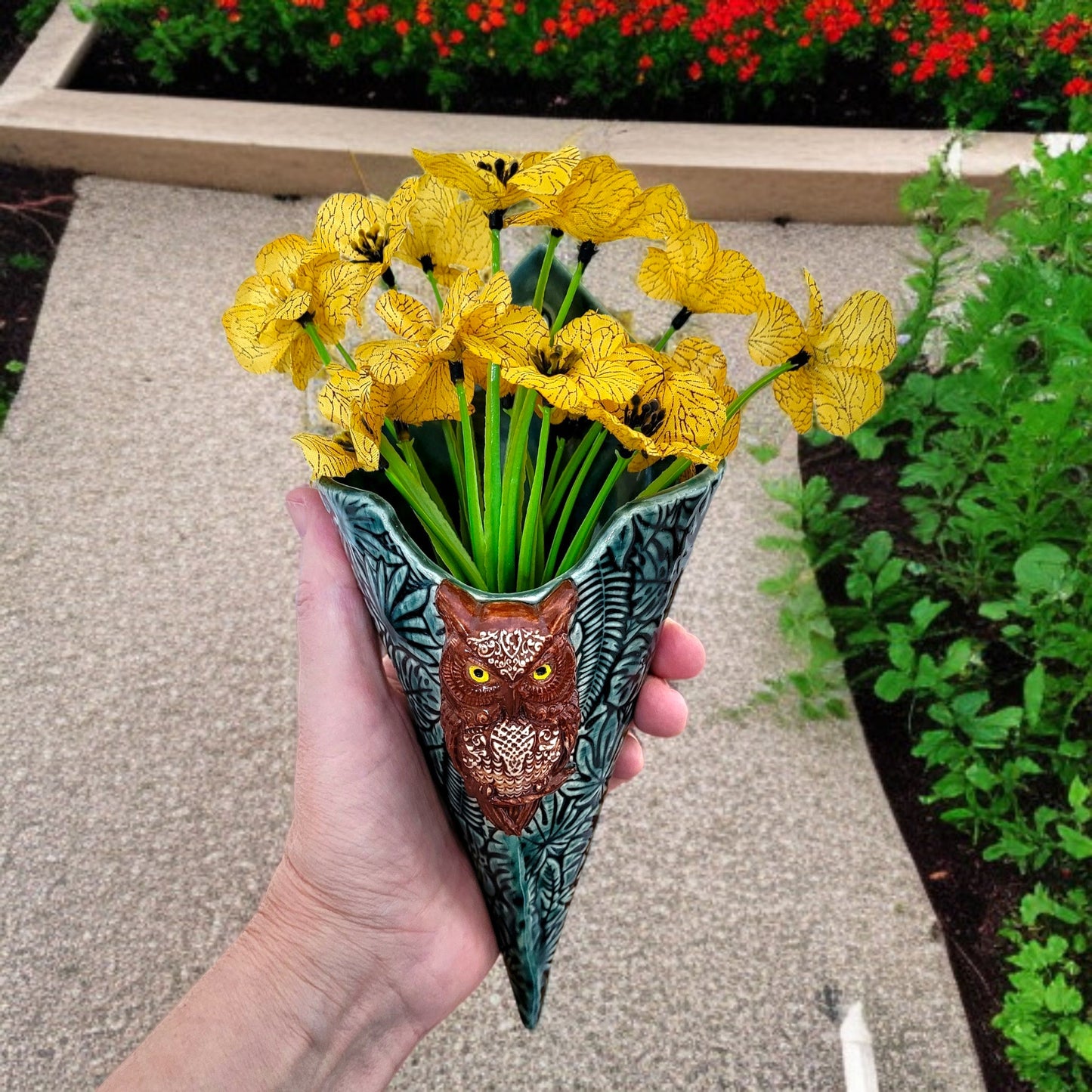 Hand holding a cone-shaped vase with yellow flowers and an owl design outdoors.