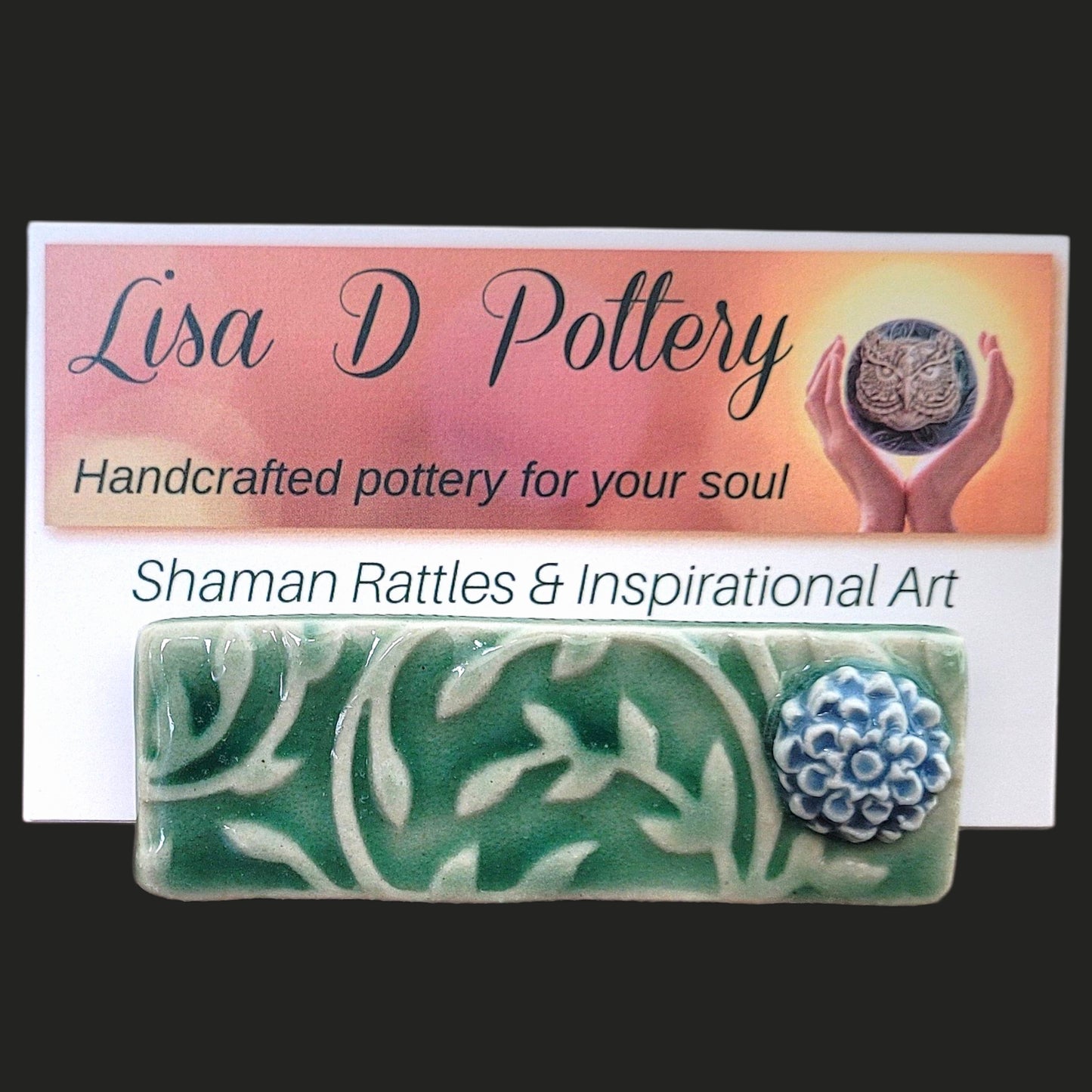 Ceramic business card holder with leaf design and blue flower holding a Lisa D Pottery business card on a gradient background