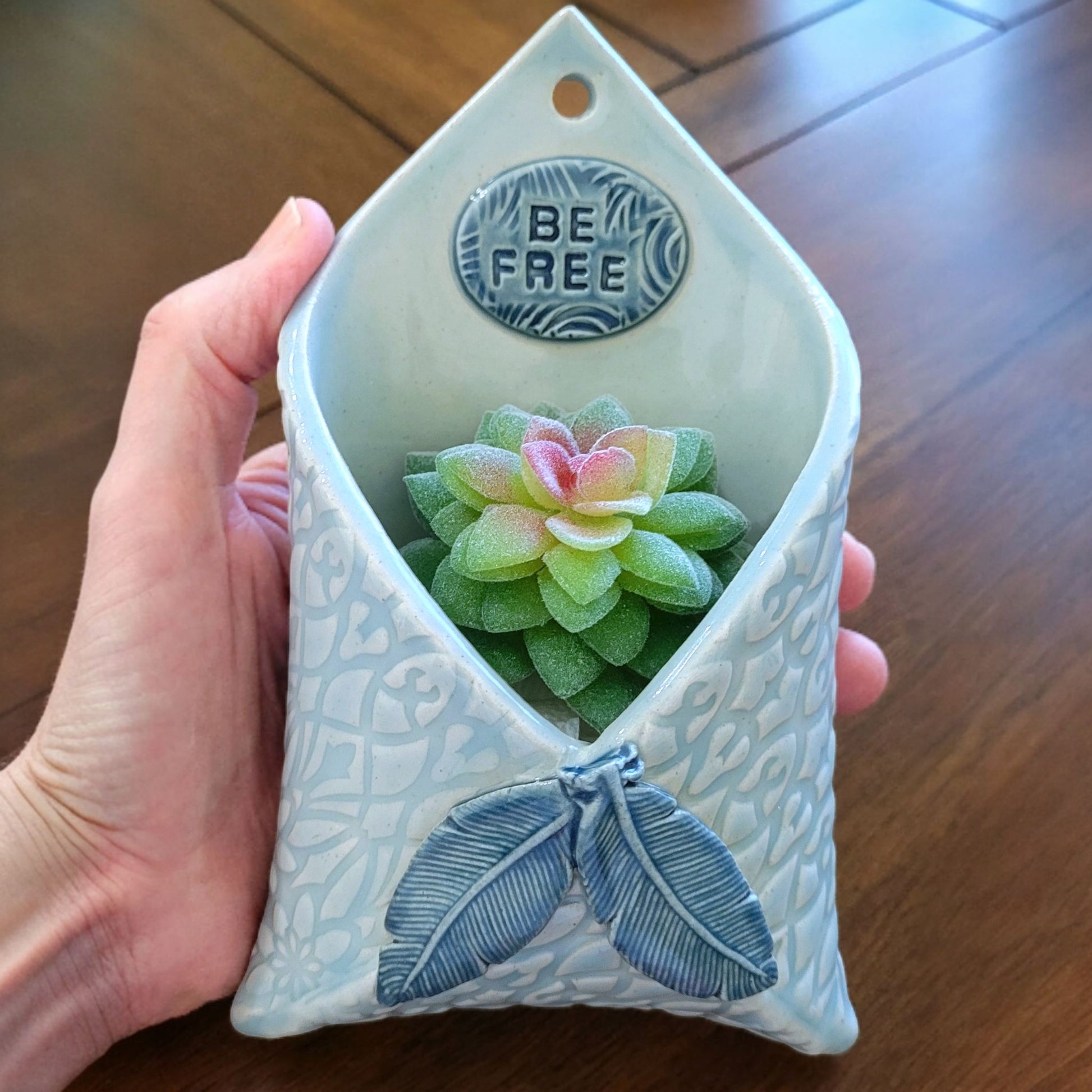 Hand holding a decorative ceramic planter with feathers and a 'Be Free' emblem.