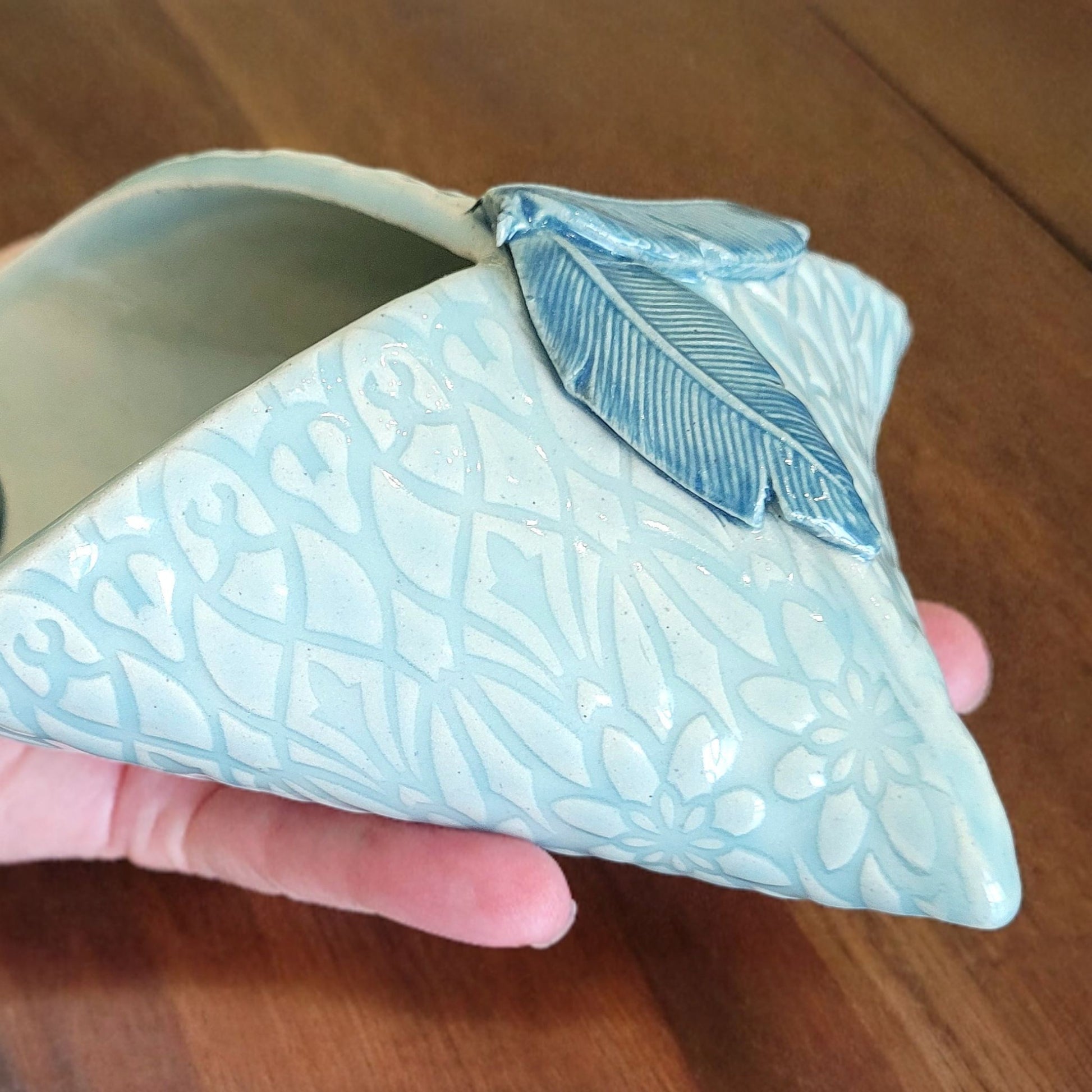 Light blue ceramic wall pocket with feather design held by a hand to show details.