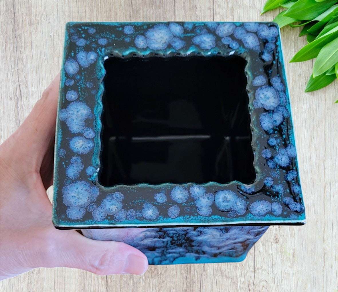 Hand holding a square ceramic planter with blue and dark gray speckled design on a light wooden surface.