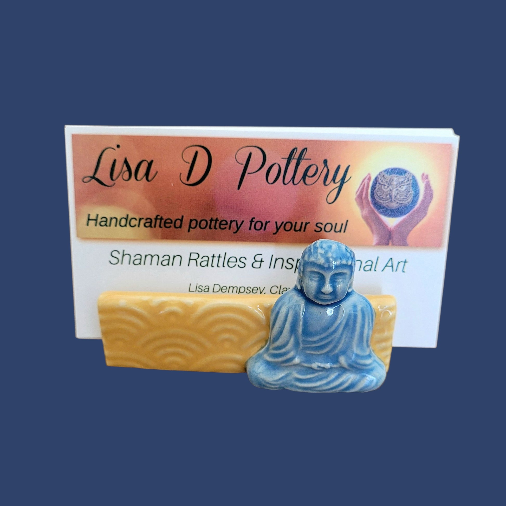 Blue ceramic Buddha figure with a business card for Lisa D Pottery on a blue background