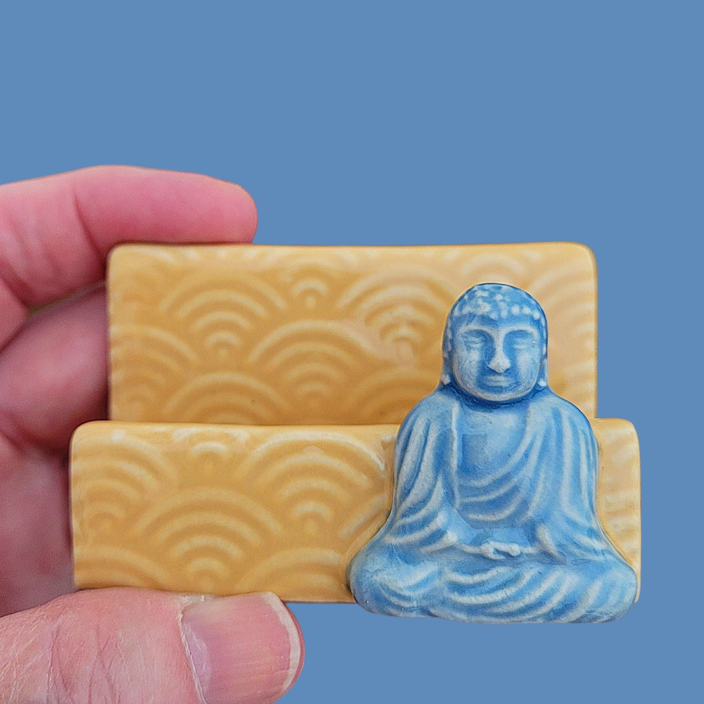 Hand holding a business card holder with a blue Buddha figurine against a yellow holder with wave patterns on a blue background
