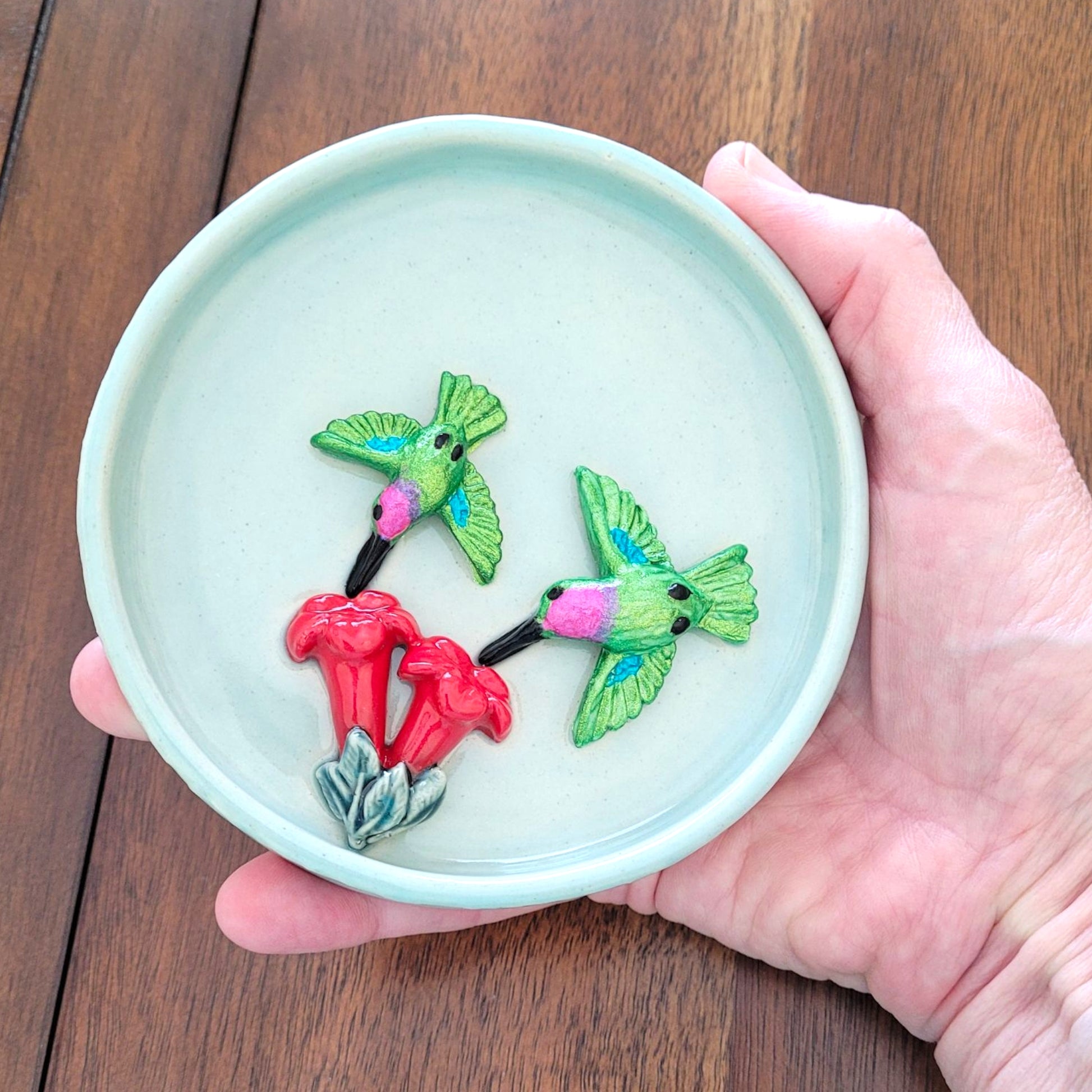 Hand holding a small round ceramic dish with  green hummingbirds and red flowers on a wooden surface.