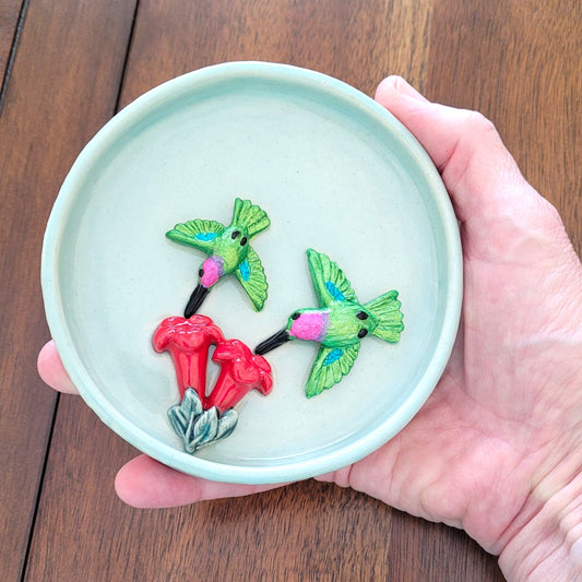 Hand holding a small round ceramic dish with  green hummingbirds and red flowers on a wooden surface.