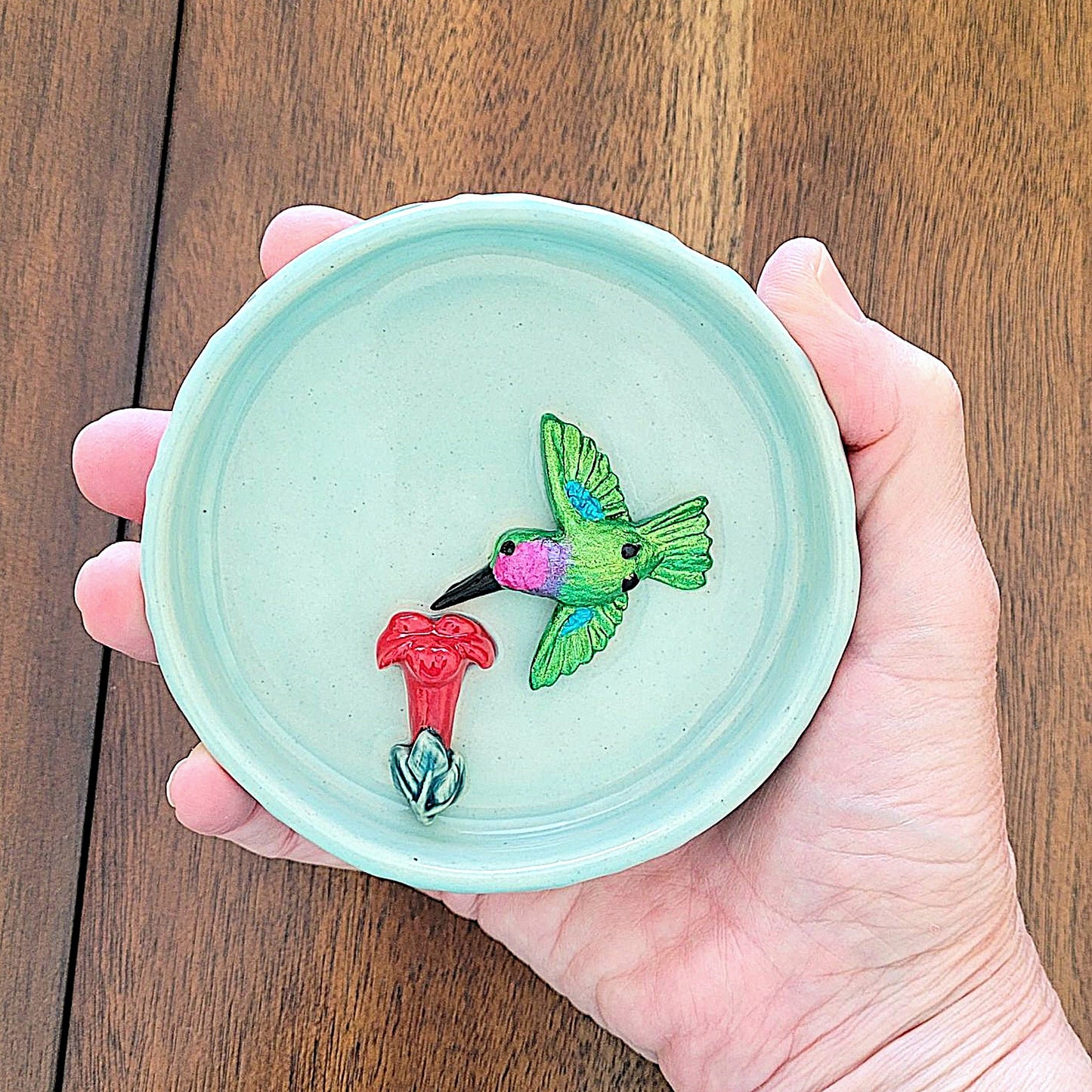 Hand holding a small ceramic dish with a green hummingbird and red flower design on a wooden surface.