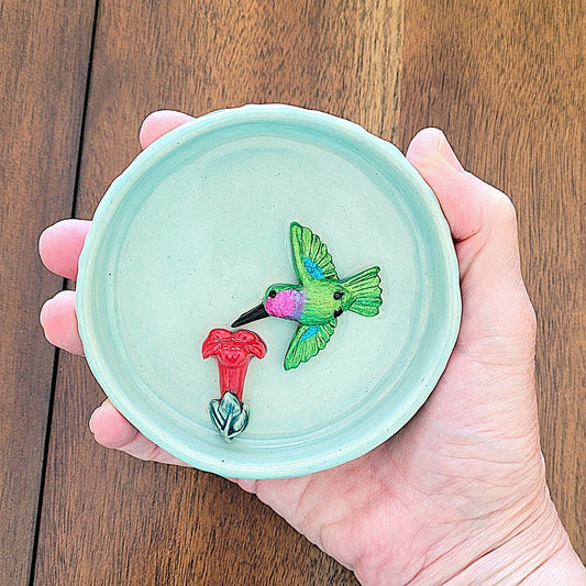 Hand holding a small ceramic dish with a green hummingbird and red flower design on a wooden surface.