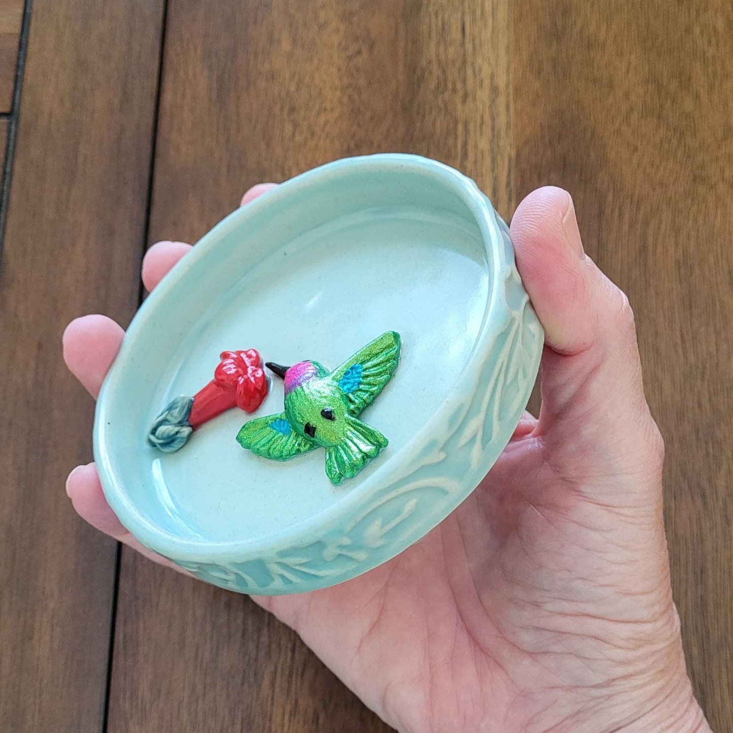 Hand holding a small blue ceramic dish with a ceramic hummingbird and red trumpet flower on a wooden surface