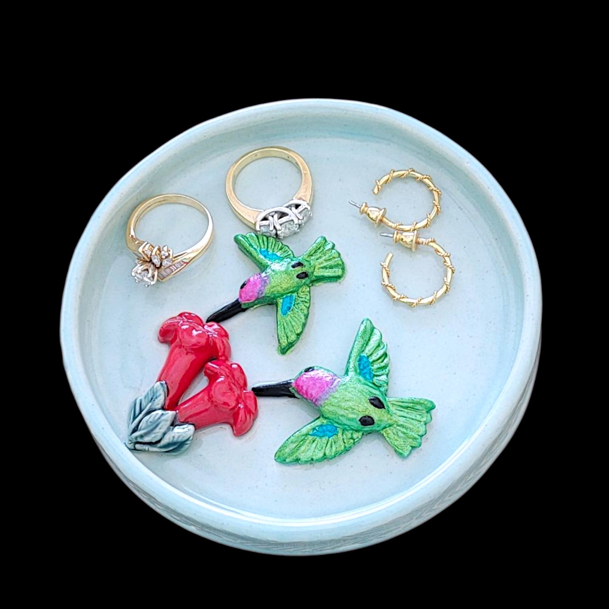 Ceramic blue dish with hummingbirds and trumpet flowers holding rings and earrings against a black background